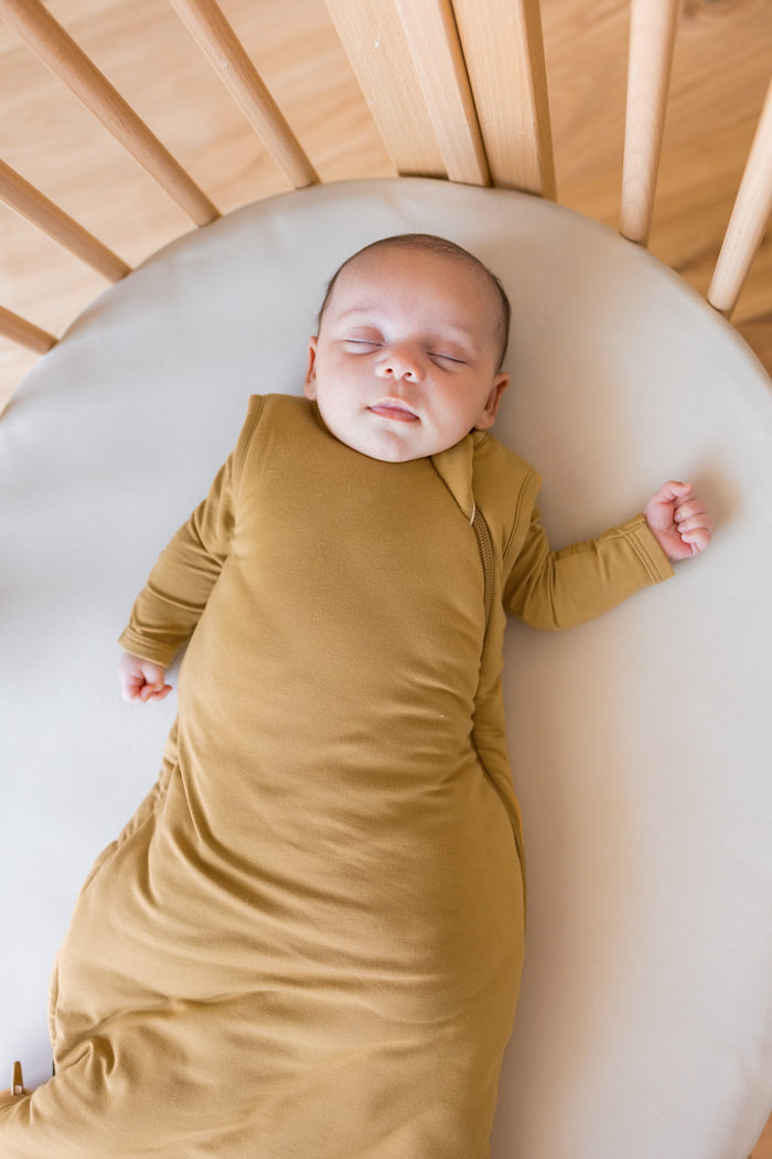 A sleeping newborn baby wearing a mustard yellow sleep sack, lying on a cream mattress inside a wooden crib.
