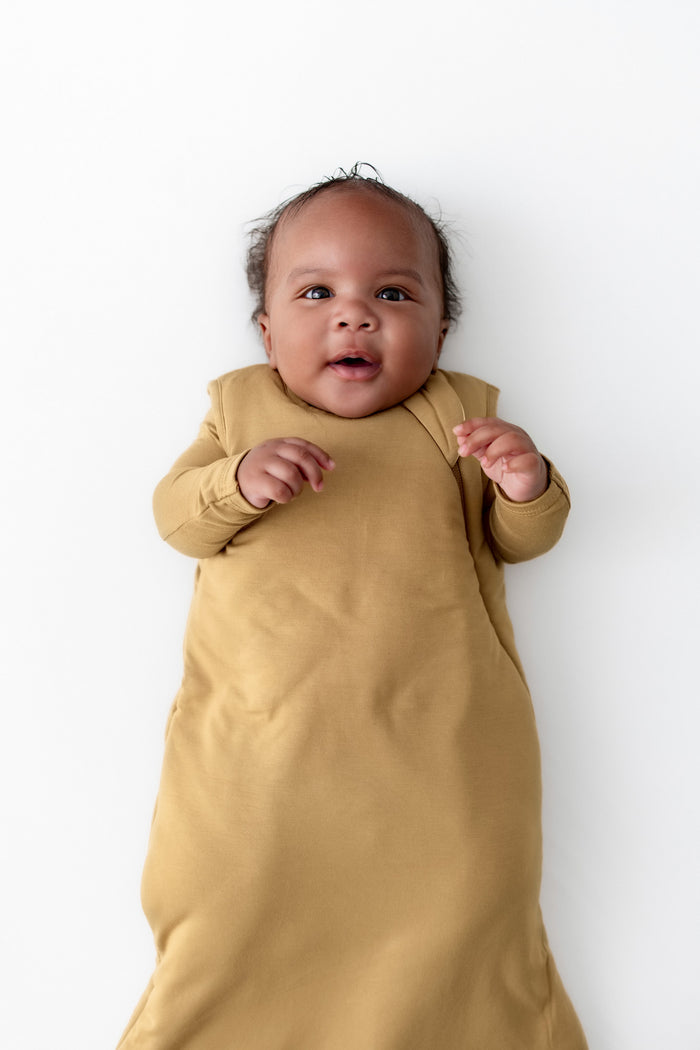A baby lying on their back wearing a mustard yellow sleep sack against a clean white background.
