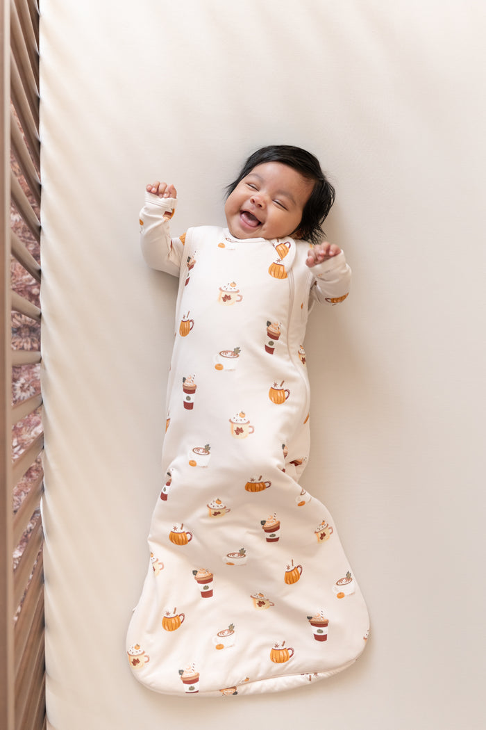 Baby smiling in Sleep Bag with Pumpkin Spice Latte 1.0 print on a beige crib sheet