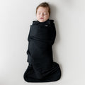 Sleeping infant laying on a light neutral blanket swaddled in the Sleep Bag Swaddler in Midnight 1.0