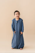 A young child wearing a slate blue zip-up sleep sack, standing against a solid beige studio background.