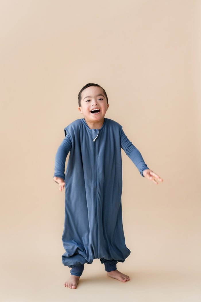 A young child wearing a slate blue zip-up sleep bag walker, standing barefoot against a solid beige studio background.