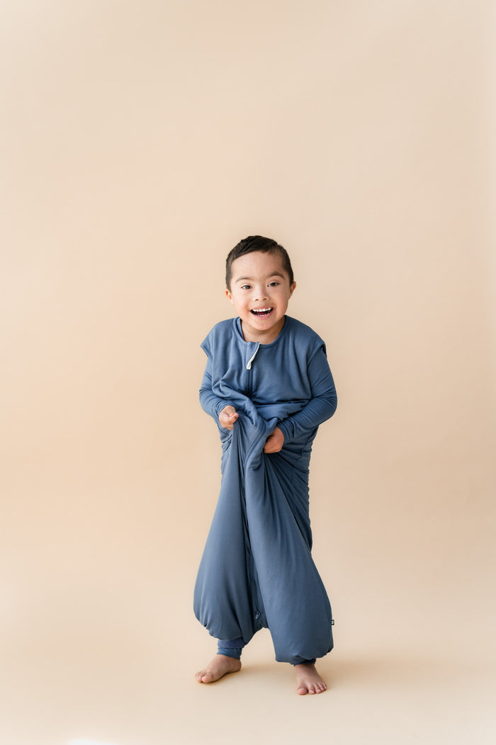 A young child stands barefoot against a plain beige studio background, wearing blue sleep sack walker