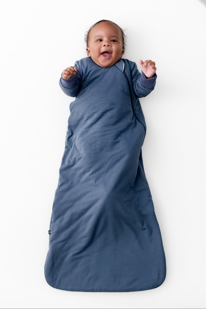 A baby lying on their back, wrapped in a slate blue long sleeve wearable blanket. The baby is smiling and has their hands raised.