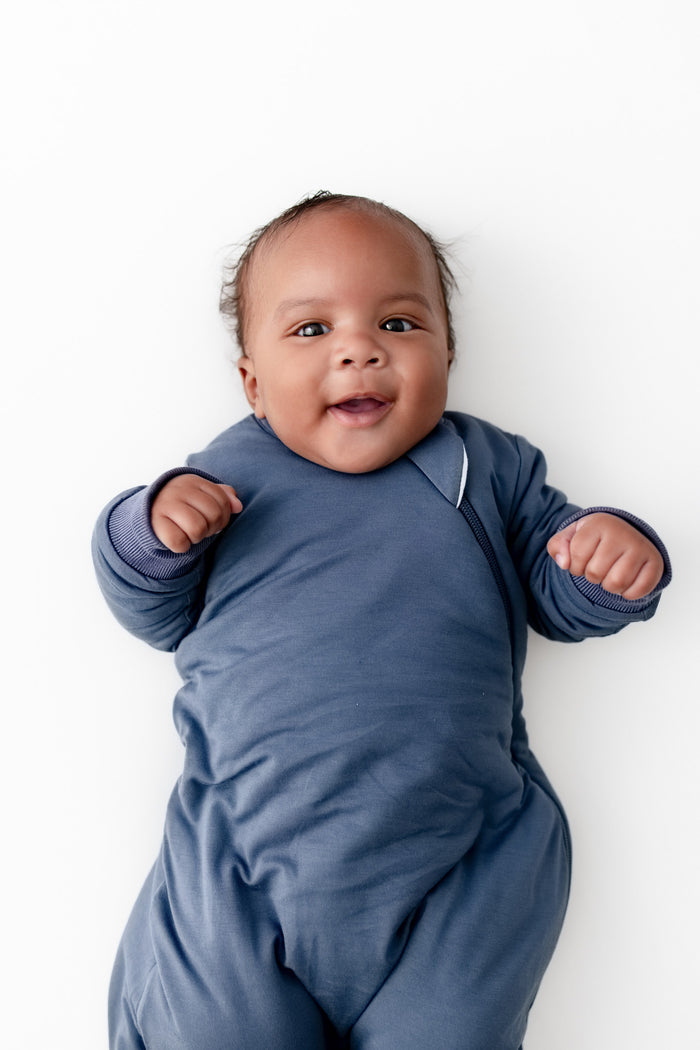 A smiling baby lying on their back wearing a slate blue long-sleeve wearable blanket against a clean white background.