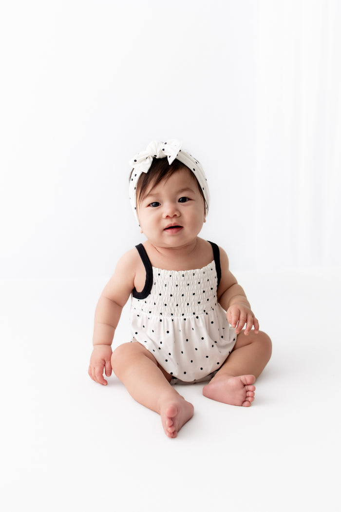 Baby wearing Midnight Polka Dot print Smocked Bubble Romper and bow headband on a white background