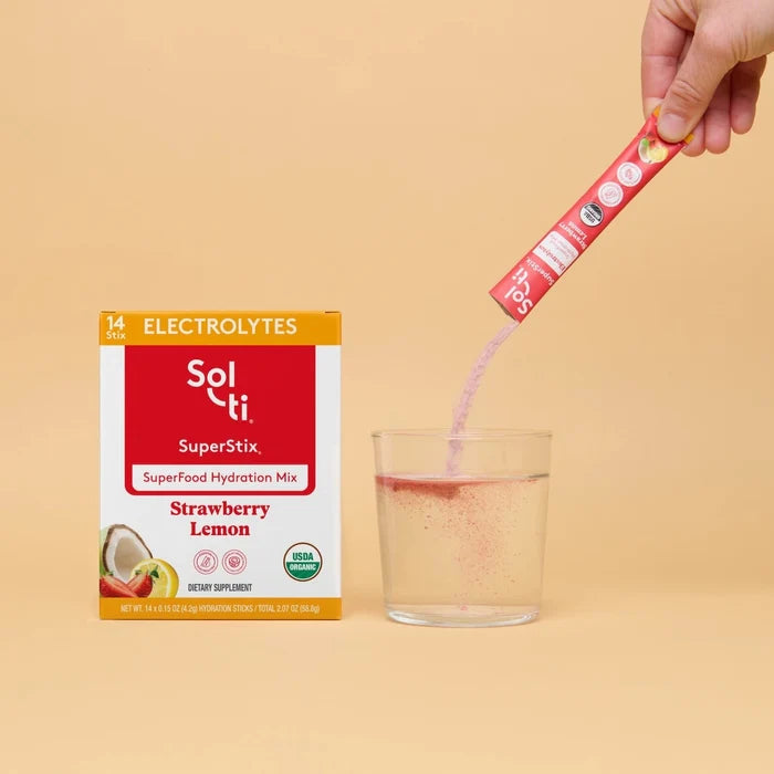 Hand pouring Sol-ti SuperStix into a glass beside the product box on a beige background