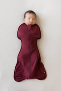 A sleeping newborn baby wrapped in a burgundy zippered swaddle bag, lying flat on a light gray background.