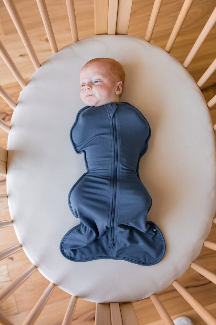A baby lying on their back inside a wooden crib, swaddled in a dark slate blue zippered swaddle bag. The baby is resting on a round cream-colored mattress pad.