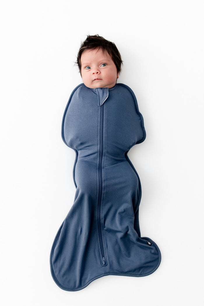 A newborn baby is wrapped in a navy blue zippered swaddle bag, lying flat on a clean white background.