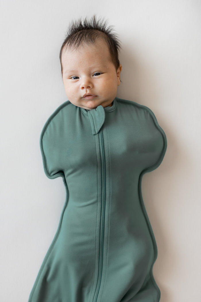 A baby wearing a sage green zip-up baby swaddle bag, lying on their back against a plain off-white wall.