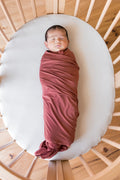 A sleeping newborn baby swaddled in a dusty terracotta  blanket, lying on a white oval mattress inside a light wood slatted crib.