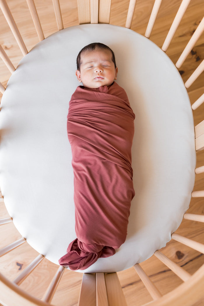 A sleeping newborn baby swaddled in a dusty terracotta  blanket, lying on a white oval mattress inside a light wood slatted crib.