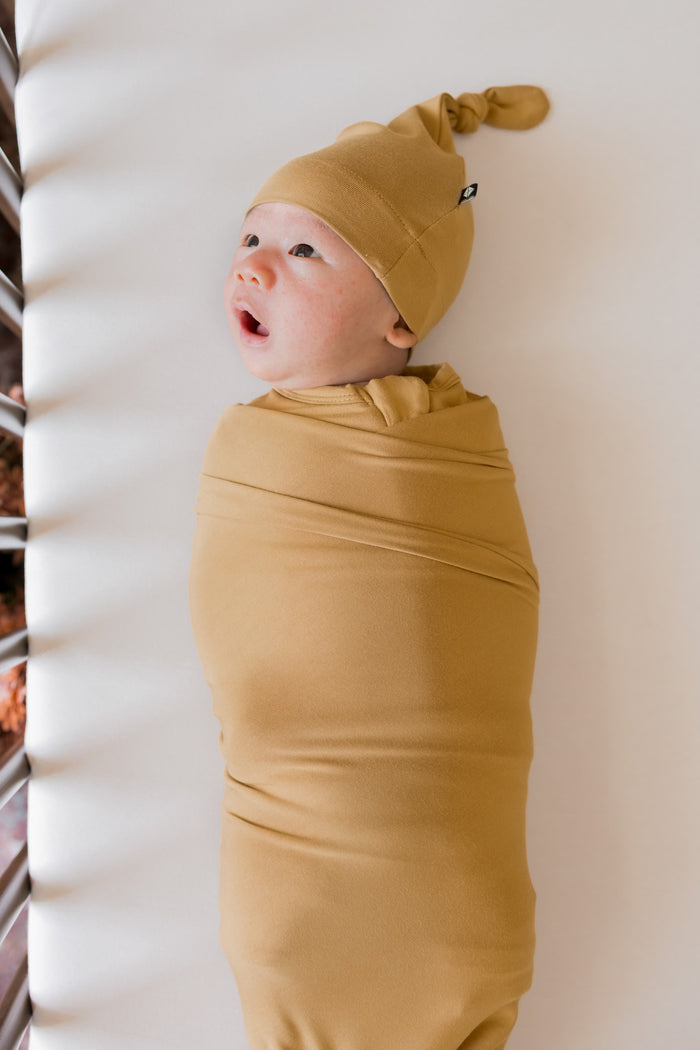 A baby swaddled in a mustard yellow blanket and wearing a matching pointed knit hat, lying on a white blanket next to a crib.