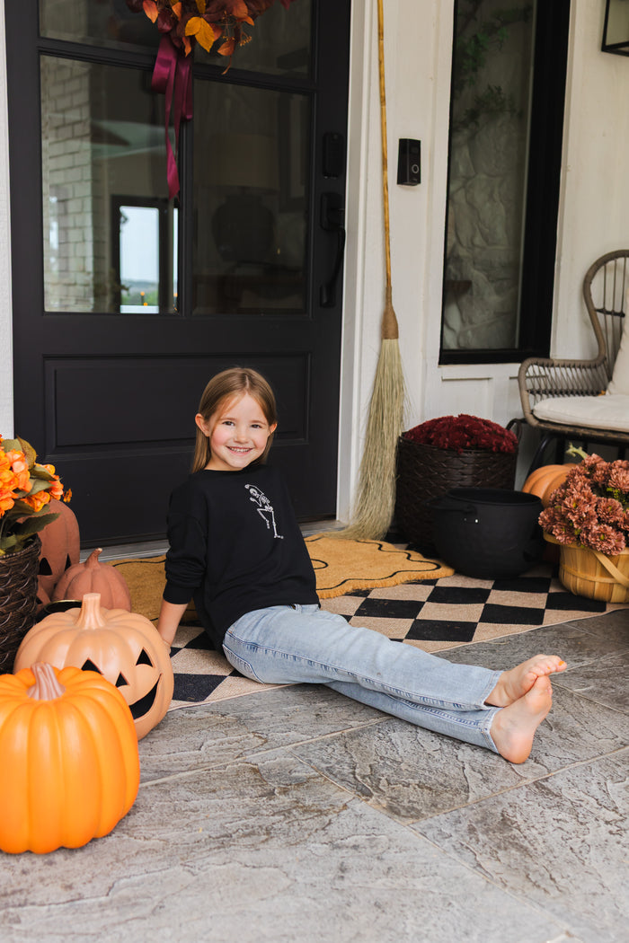Girl smiling in Black Sweatshirt with Skeleton print in the center sitting outdoors with Halloween decorations