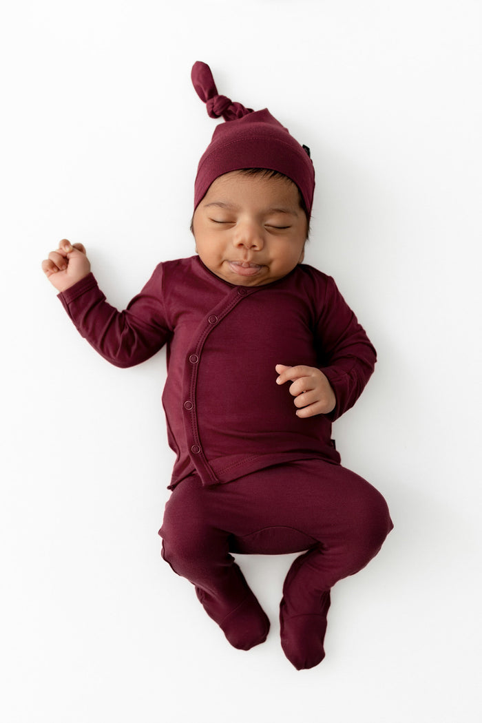 A sleeping newborn baby wearing a matching burgundy long-sleeve footed take me home set and knotted beanie hat, laid flat on a clean white background.