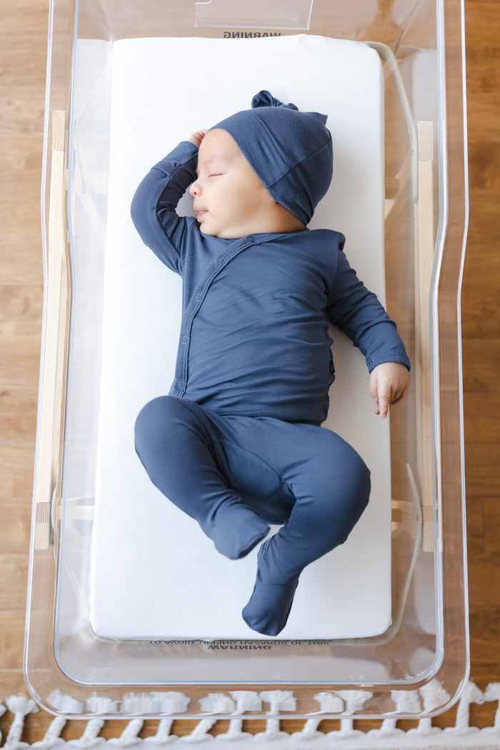 A sleeping newborn baby wearing a navy blue long-sleeve footed take me home set and matching beanie hat, lying on a white mattress inside a clear plastic bassinet.