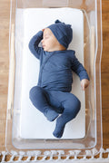 A sleeping newborn baby wearing a navy blue long-sleeve footed take me home set and matching beanie hat, lying on a white mattress inside a clear plastic bassinet.