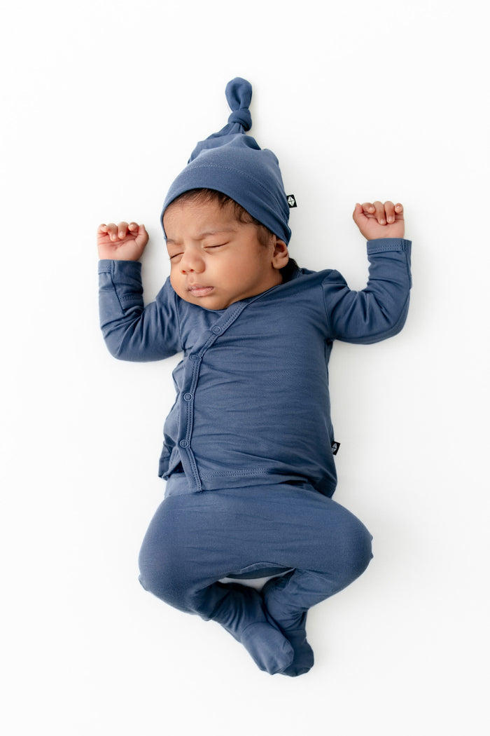 A sleeping newborn baby wearing a matching slate blue long-sleeve outfit with a pointed knit hat, photographed from above on a clean white background.