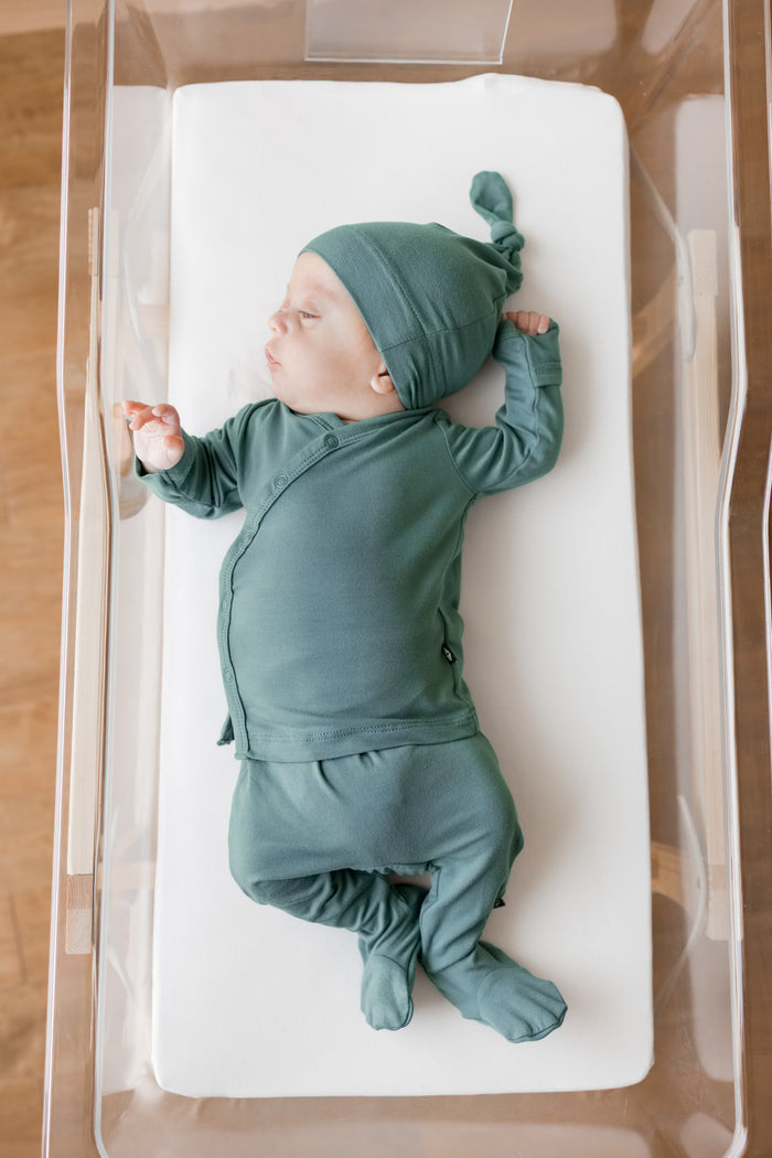 A sleeping newborn baby wearing a matching sage green footed take me home set and knit hat, lying on a white mattress inside a clear plastic bassinet.