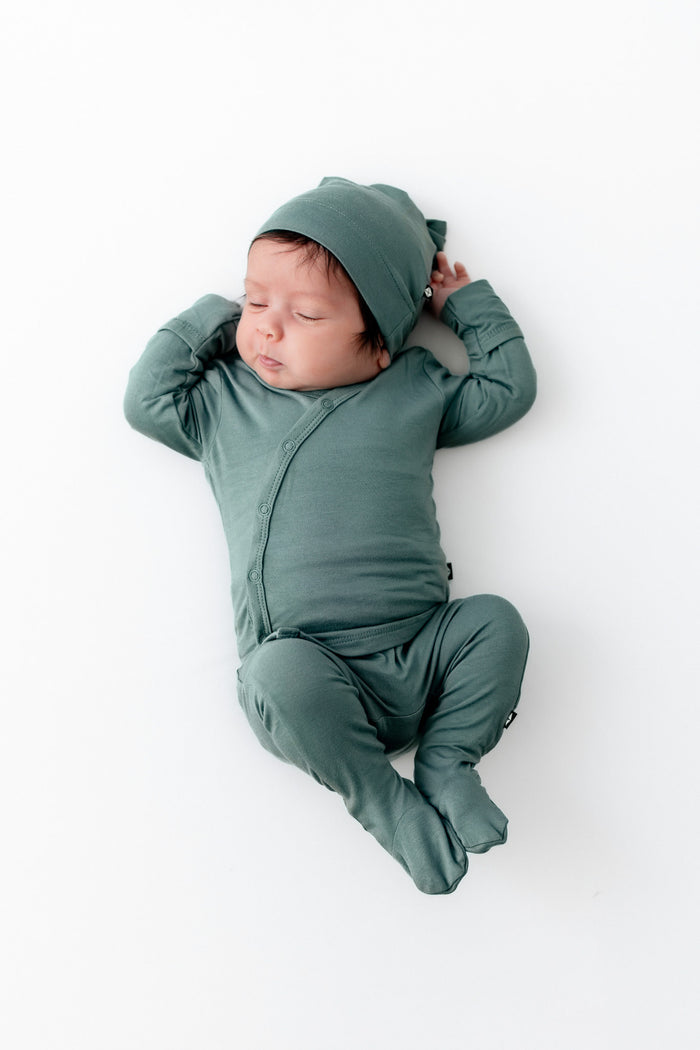 A sleeping newborn baby wearing a matching sage green footed take me home set and cap, lying on their back against a clean white background.