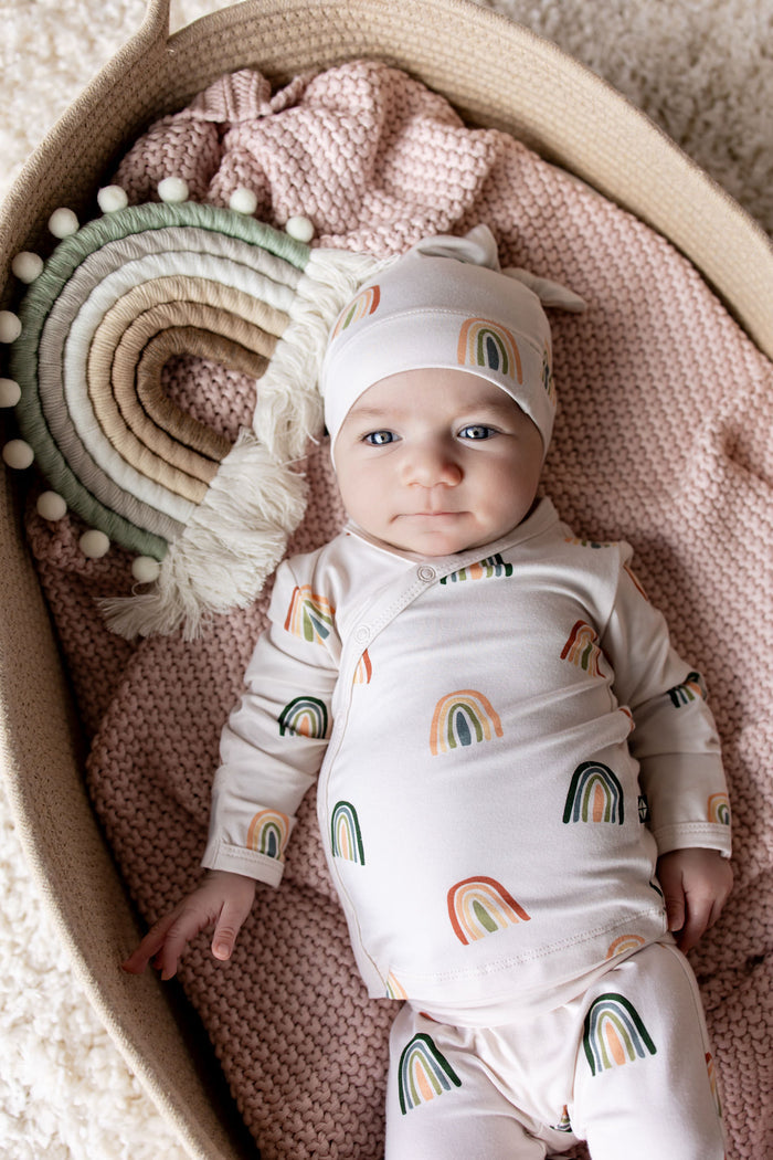 Baby in Take Me Home Set with Cap in Retro Rainbow print lying in a basket over a pink blanket with a retro rainbow prop