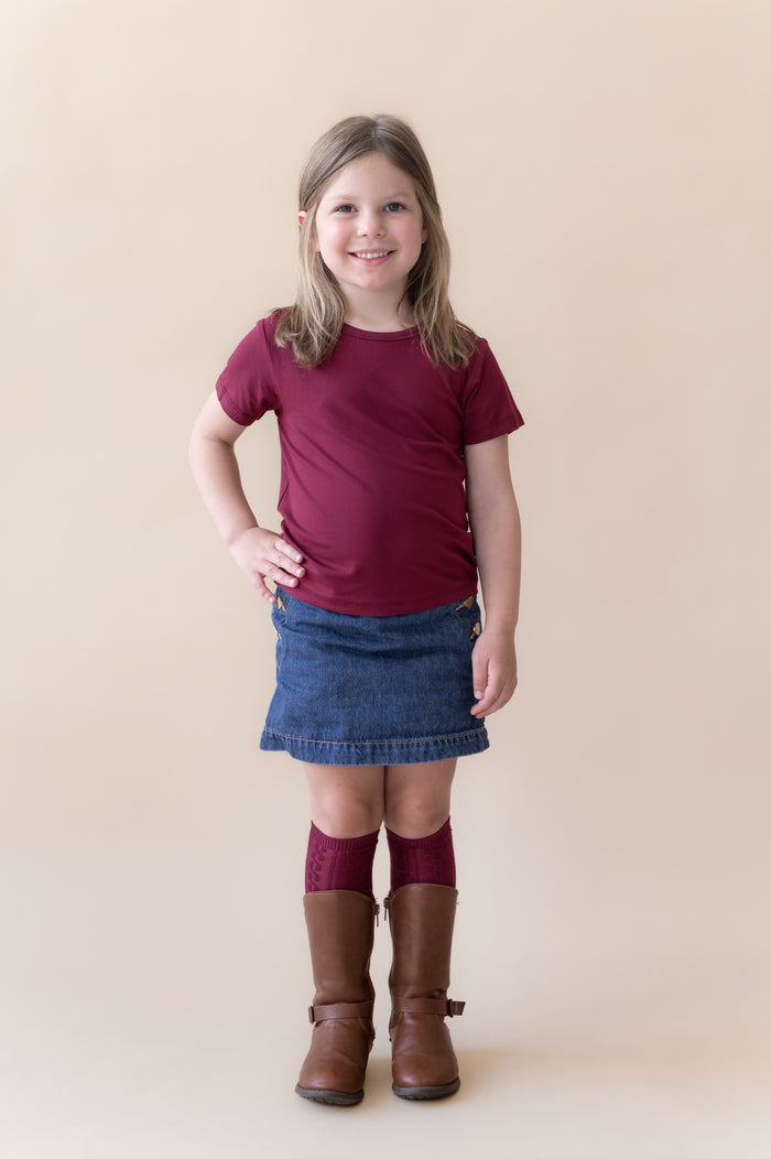 Girl posing Toddler Basic Tee in Burgundy on blue skirt and burgundy knee high socks and boots on a beige background