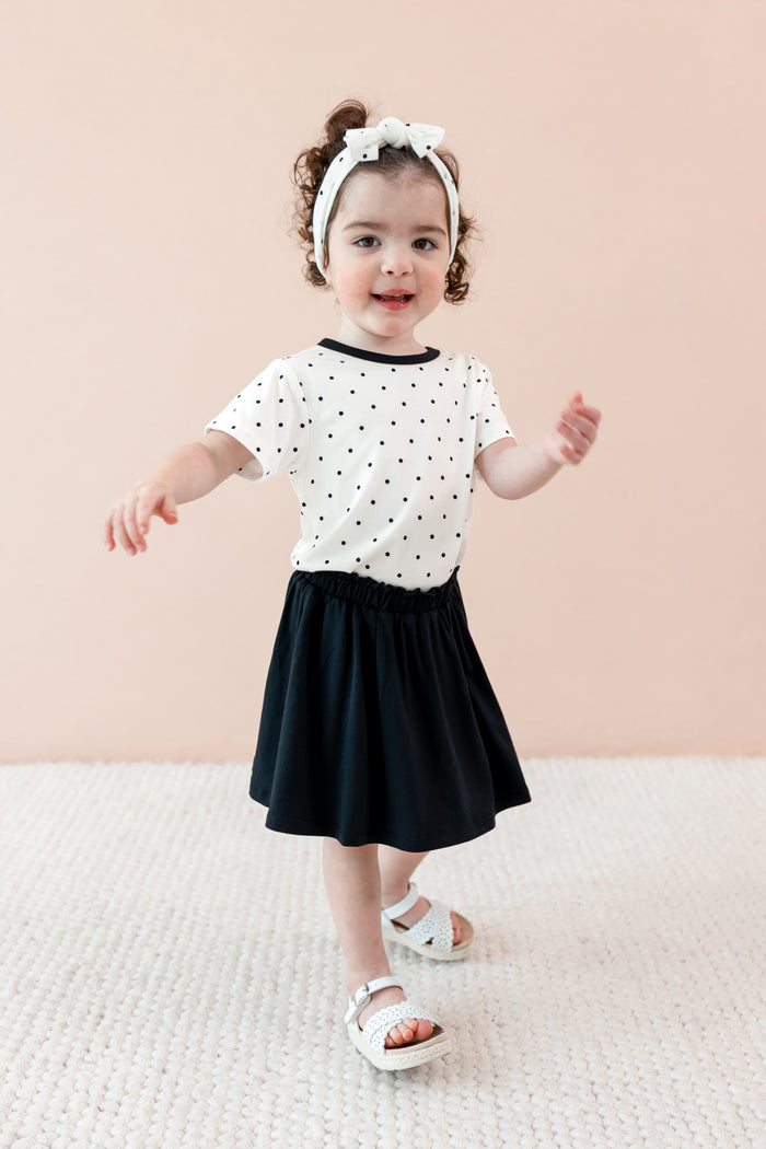 Child wearing Midnight Polka Dot print Toddler Basic Tee and bow headband over a black skort on a beige background