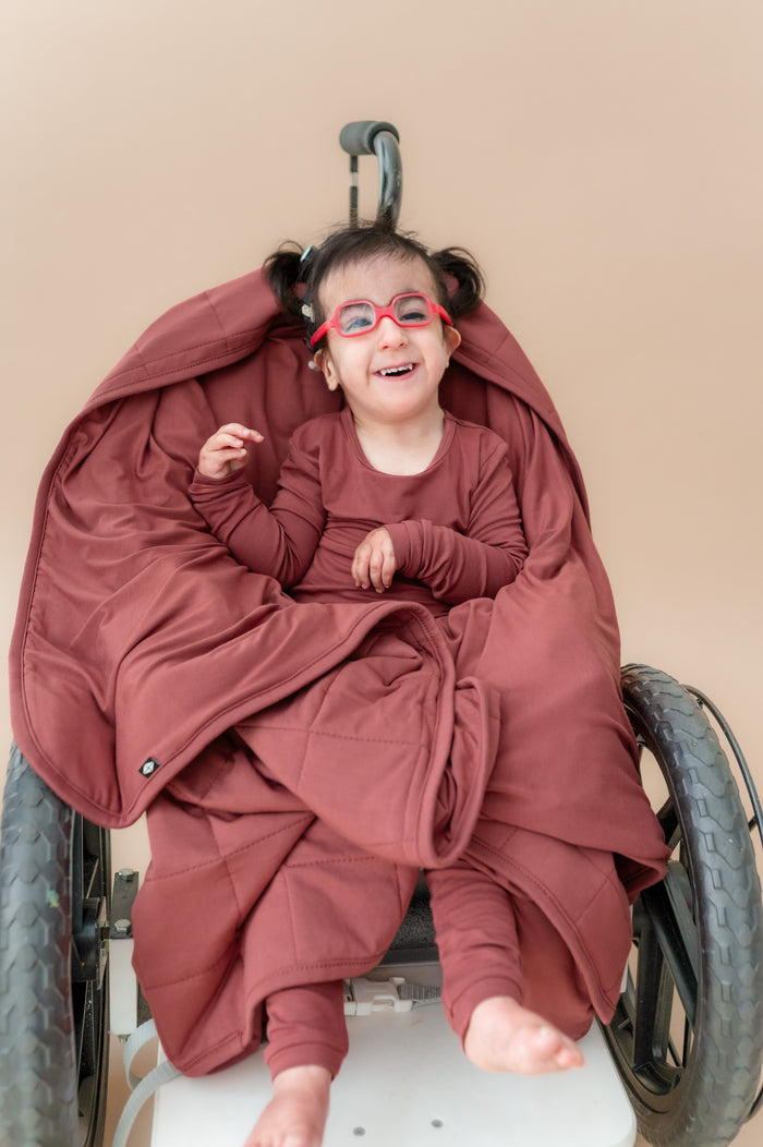 Child sitting on a wheel chair wrapped in Toddler Blanket in Ember 2.5 wearing a similar color long sleeve pajamas