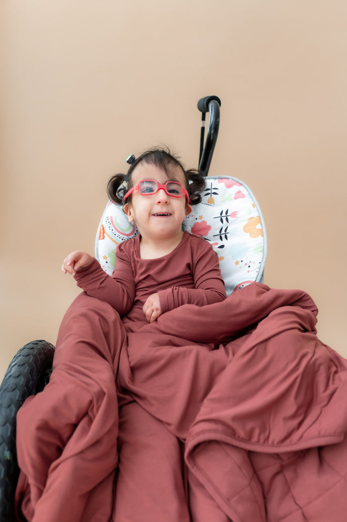 Child sitting on a wheel chair wrapped in Toddler Blanket in Ember 2.5 wearing a similar color long sleeve pajamas