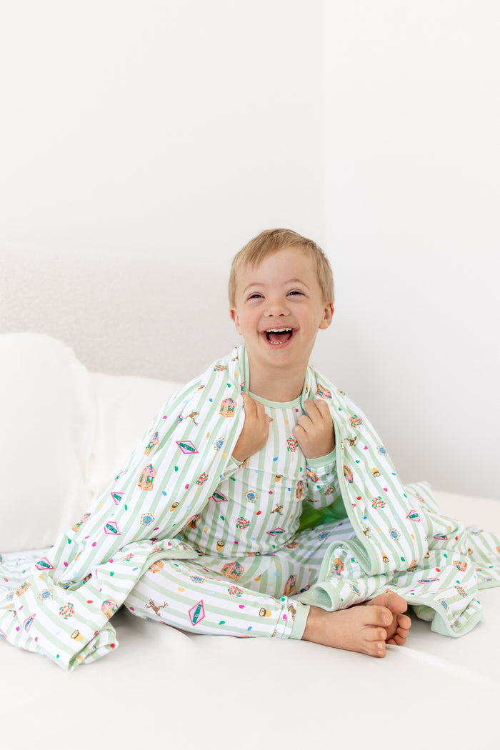 Child sitting on a white bed wrapped in a green and white striped toddler blanket with colorful Harry Potter patterns wearing a similar print long sleeve pajama set