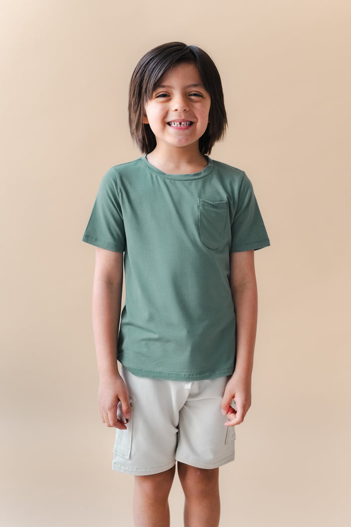 Boy wearing Toddler Crew Neck Tee in Pine over white shorts on a beige background