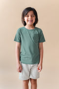 Boy wearing Toddler Crew Neck Tee in Pine over white shorts on a beige background
