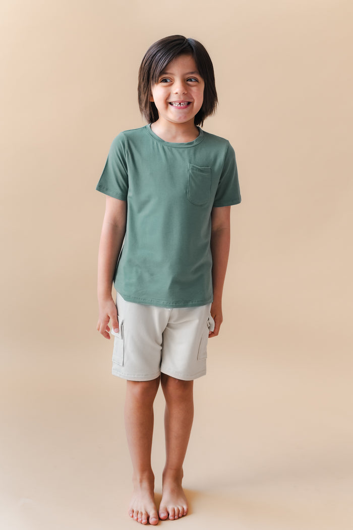 Boy smiling in Toddler Crew Neck Tee in Pine over white shorts on a beige background
