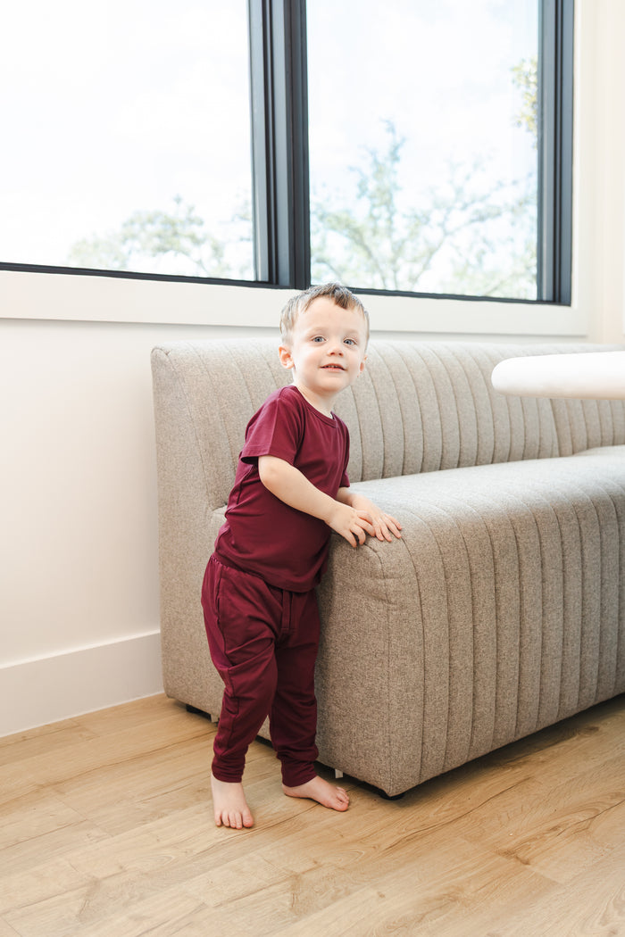 Child wearing Toddler Jogger Pant in Burgundy under Toddler basic tee in burgundy standing against a sofa