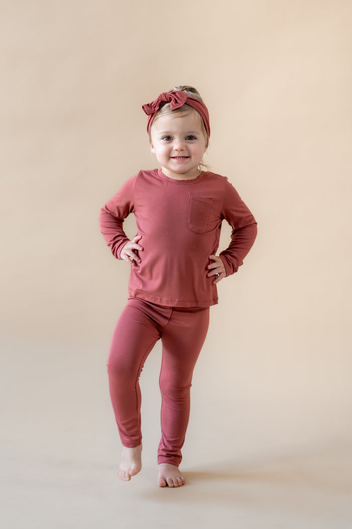 Girl wearing Toddler Leggings in Ember over similar color Toddler Crew Neck Tee and knotted bow headband on a beige background