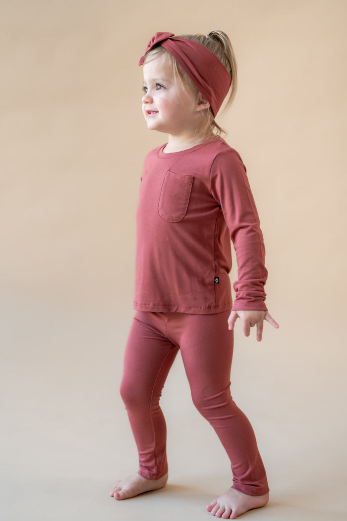 Girl posing Toddler Leggings in Ember over similar color Toddler Crew Neck Tee and knotted bow headband on a beige background