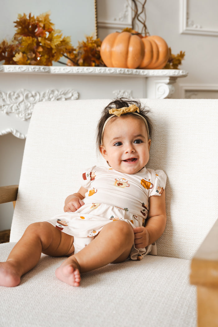 A baby girl sitting on a white upholstered chair, wearing a cream wrap dress with a pumpkin spice latte print and a gold headband. The background features a white fireplace mantel decorated with an orange pumpkin and autumn leaves.