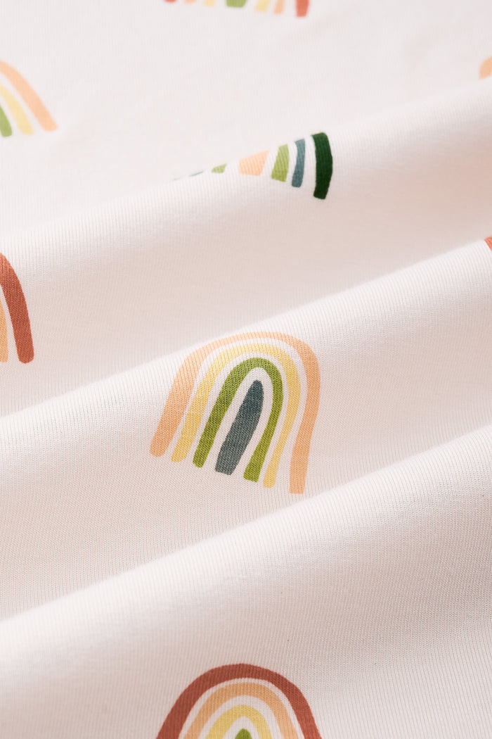 An all-over print featuring alternating rows of boho style rainbows in alternating directions over a light beige background color called Vanilla. 

One row of rainbows has an outermost arc colored in a dark cool green, followed by a cool sage green, then a light olive green, and finished with an inner core colored in a light apricot. 

Another row of of rainbows has an outermost arc colored in a burnt orange called Apricot, followed by a light peach, then a light yellow called Lilikoi, and finished with an