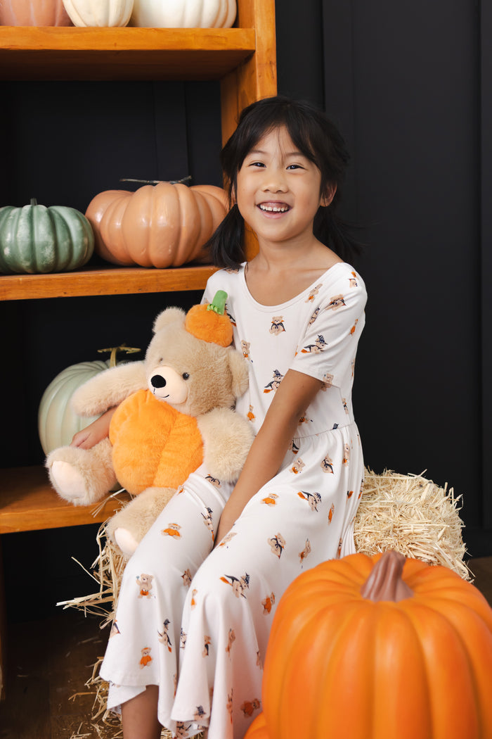 Girl sitting with a stuffed teddy bear wearing Twirl Dress in Halloween Teddy print with pumpkin decorations around