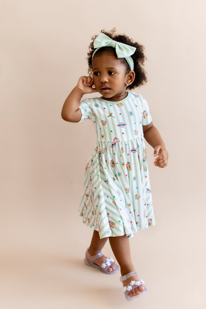 Child wearing a green and white striped twirl dress with colorful Harry Potter patterns and a green bow headband on a beige background