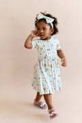Child wearing a green and white striped twirl dress with colorful Harry Potter patterns and a green bow headband on a beige background