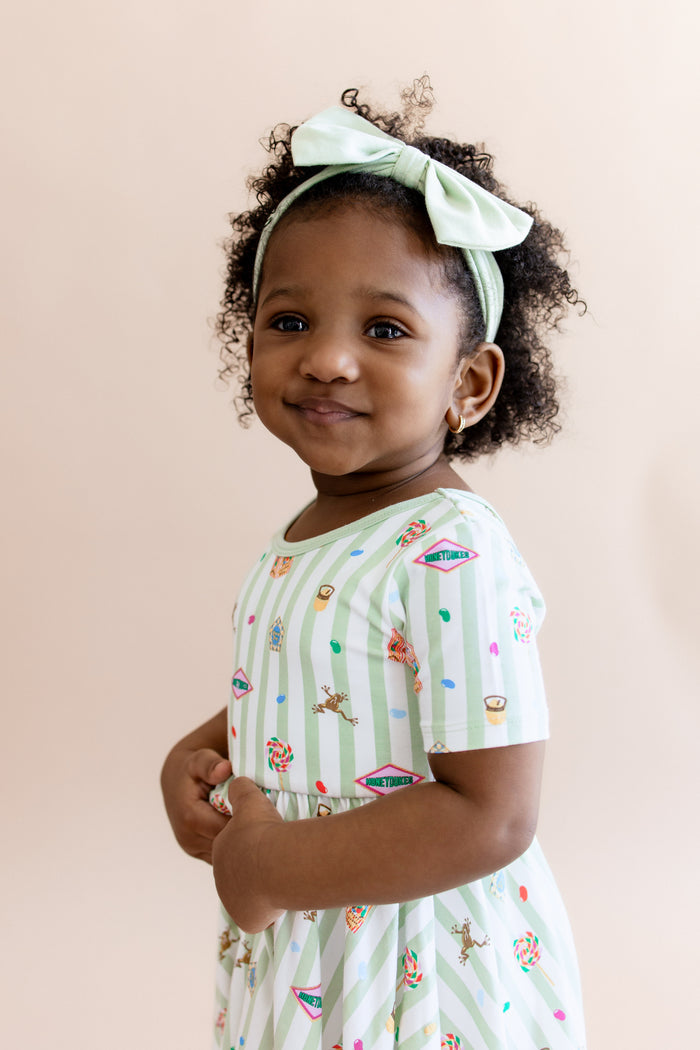 Child in a twirl dress having green and white stripes fabric with colorful Harry Potter patterns and a green headband on a beige background