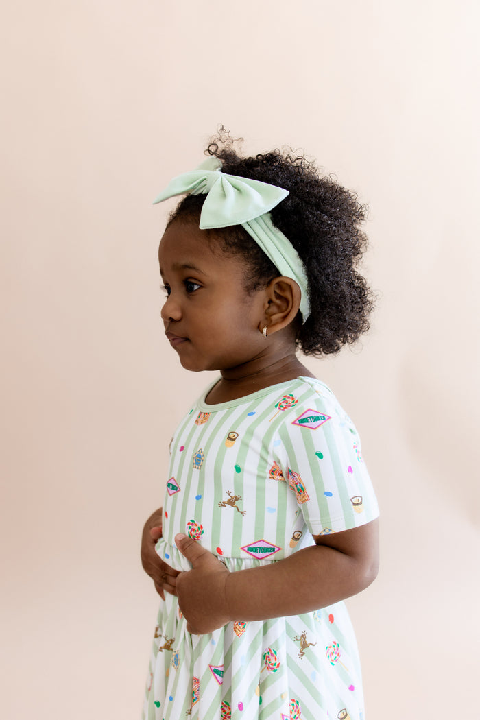 Side view of child in a green and white striped twirl dress with colorful Harry Potter patterns and a green bow headband on a beige background