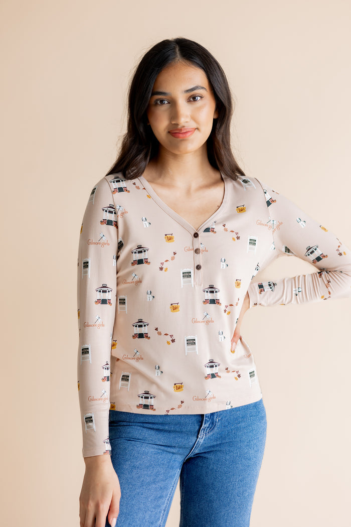 A woman modeling a long-sleeve henley style top with a  Gilmore Girls inspired print, paired with blue jeans, against a solid beige background.