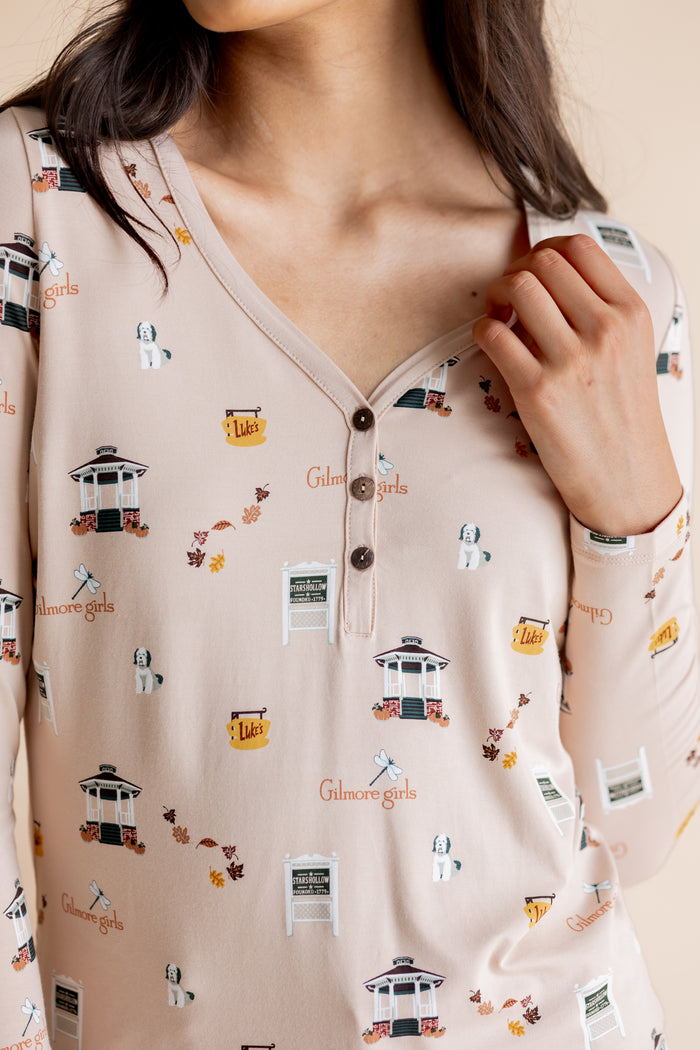 A close-up of a woman wearing a long-sleeve henley top in a light blush pink color, decorated with a whimsical print of gazebos, fall leaves, and the word 'Gilmore Girls'.