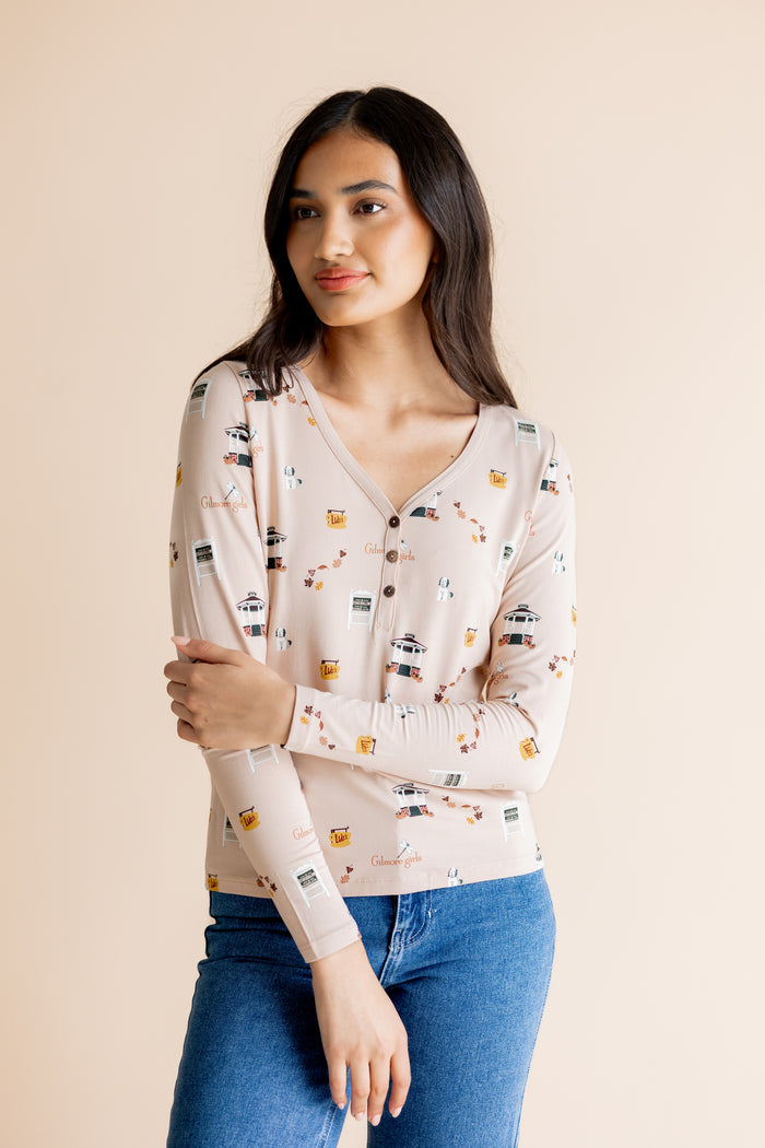 A woman modeling a light beige long-sleeve henley top with a  Gilmore Girls inspired print, paired with blue jeans, against a solid beige background.