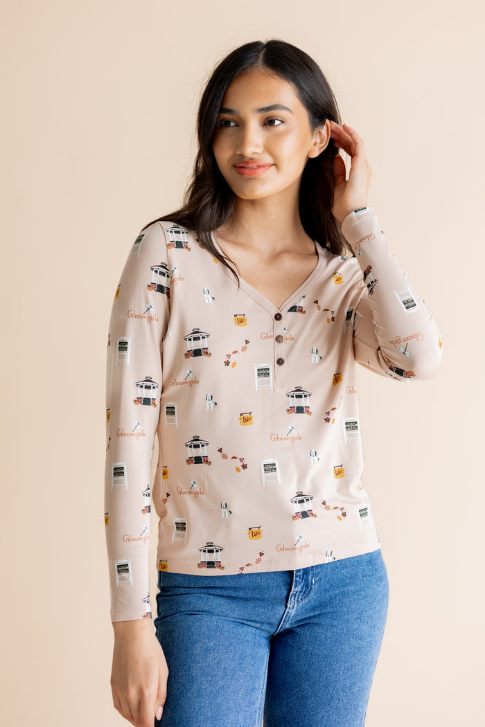 A woman modeling a long-sleeve henley top with a beige base and a Gilmore Girls inspired print, paired with blue jeans against a solid beige background.