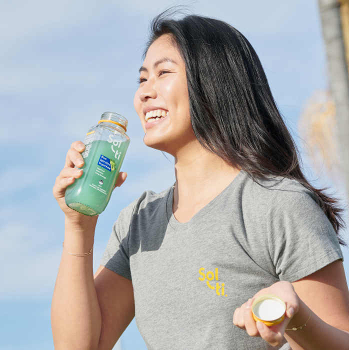 Person smiling, holding a Sol-ti Blue Spirulina SuperAde glas bottle, wearing a gray cotton tee against blue background