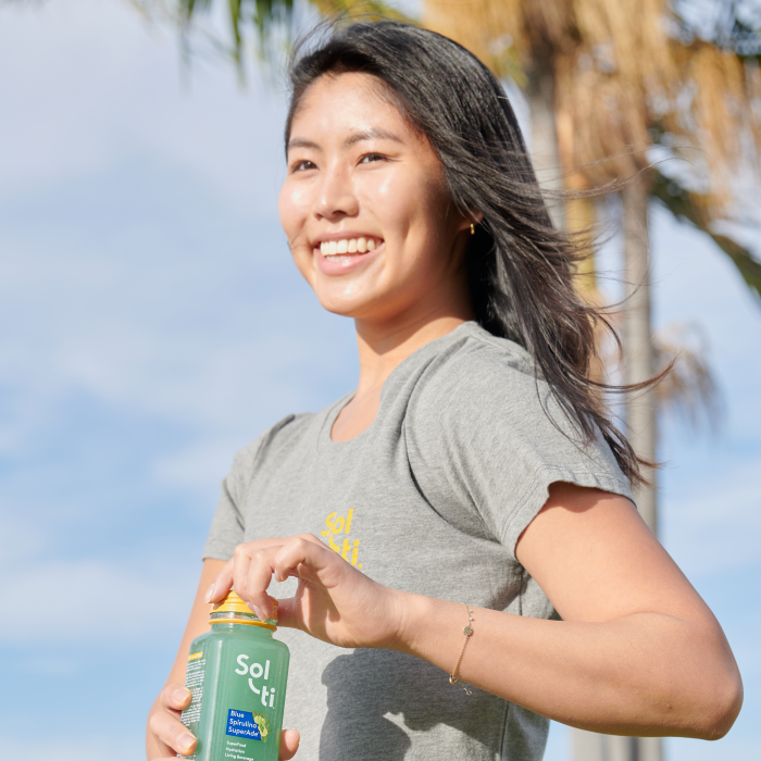 Person smiling, holding a Sol-ti Blue Spirulina SuperAde glas bottle, wearing a gray cotton tee against blue background with palm trees